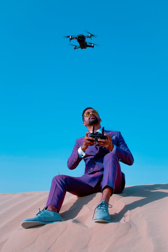 services-04 A stylish man in a purple suit controls a drone in the Dubai desert under a clear blue sky.
